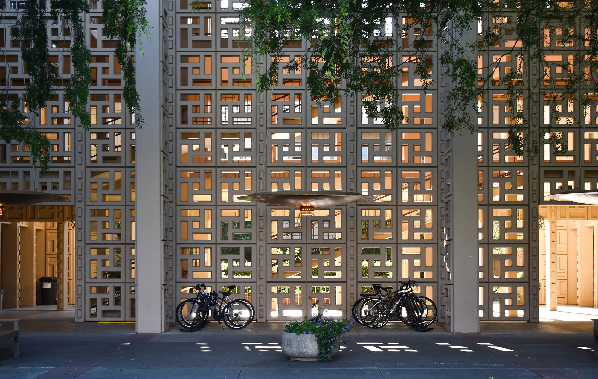 A wall of pale concrete screen block between tall square columns, with sunlight and green foliage showing through the openings and two parked bicycles at its base.