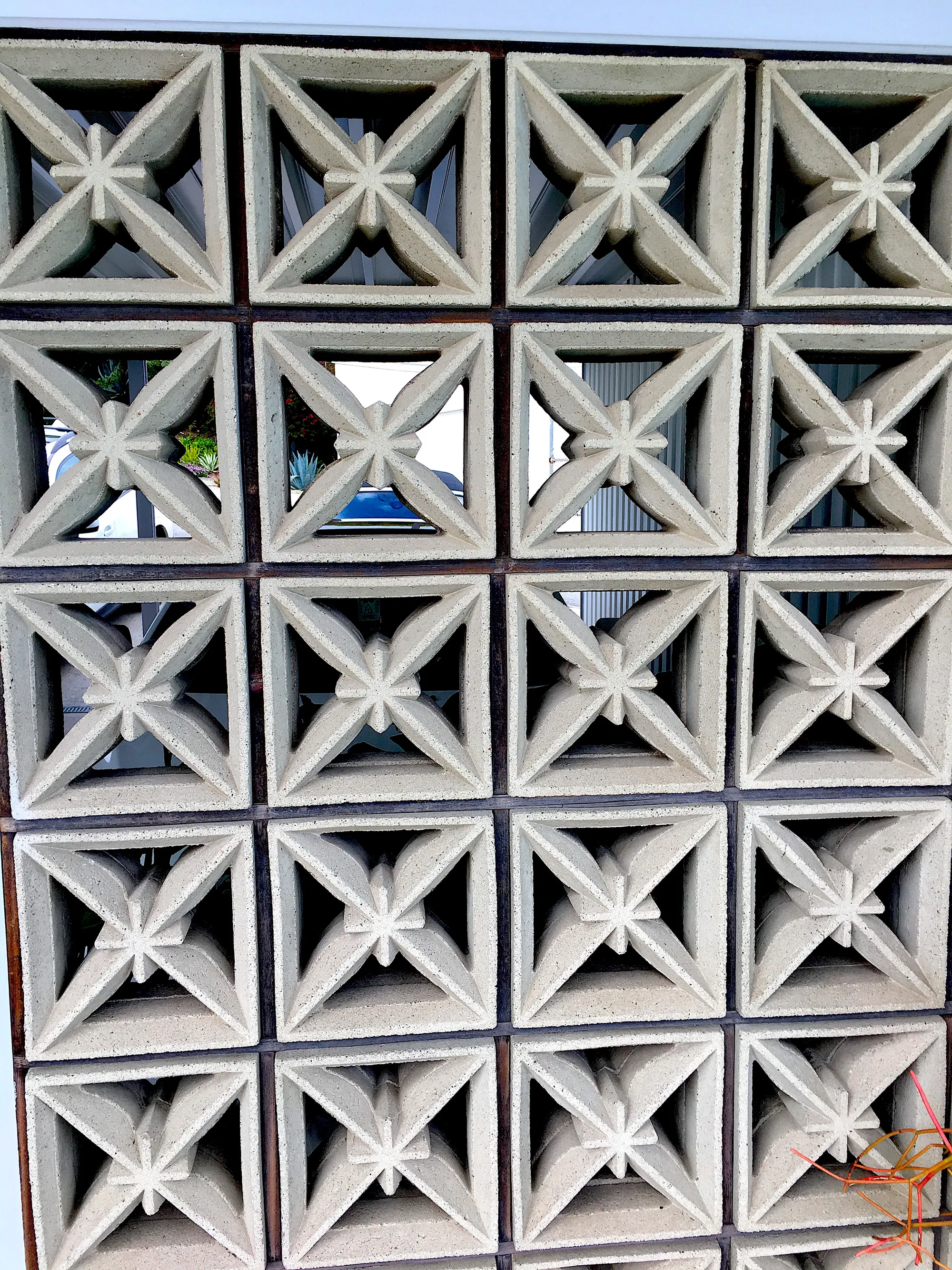Rows of square concrete screen blocks, each pierced with an X-shaped starburst pattern, held in a dark metal grid with a car and plants visible through the openings.