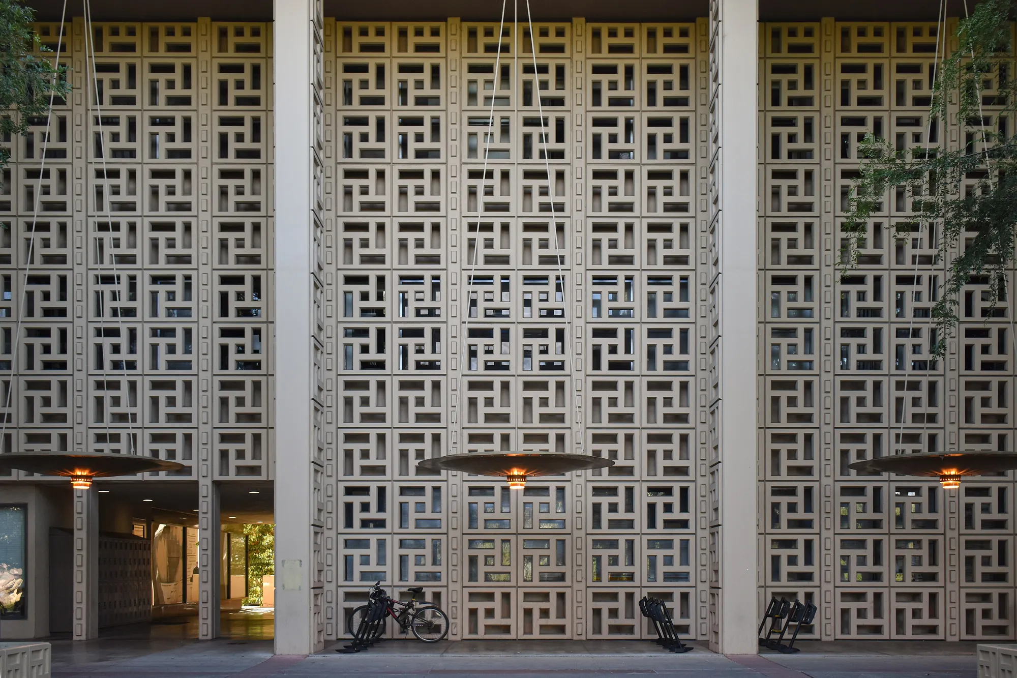 A broad facade of square concrete screen blocks in a repeating geometric pattern, divided by tall columns, with round lamp shades hanging in front of it.