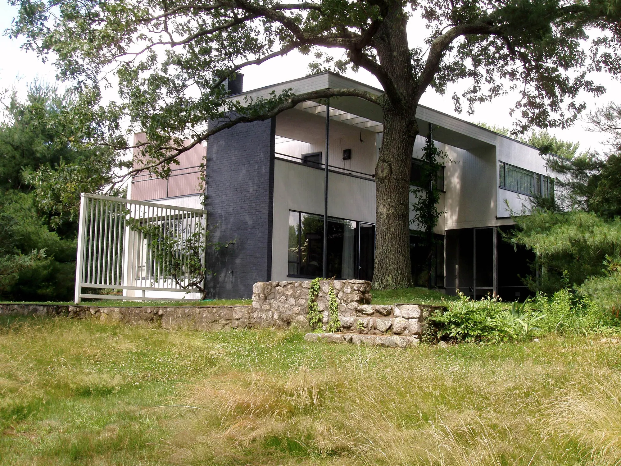 A white and dark-brick house seen from the side rear, with a fieldstone wall in front of it.