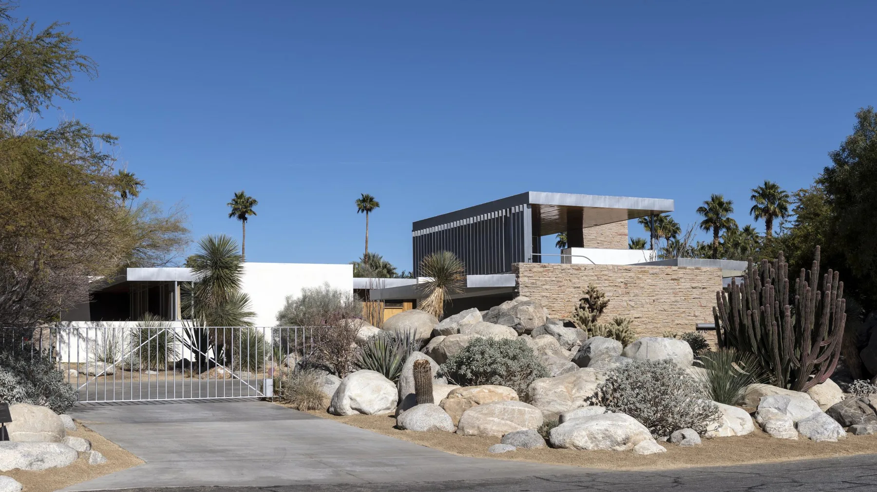 The Kaufmann Desert House seen from the street behind a metal gate, with boulders, cactus, a stone wall, an upper pavilion with a louvered fascia, palms and a blue sky.