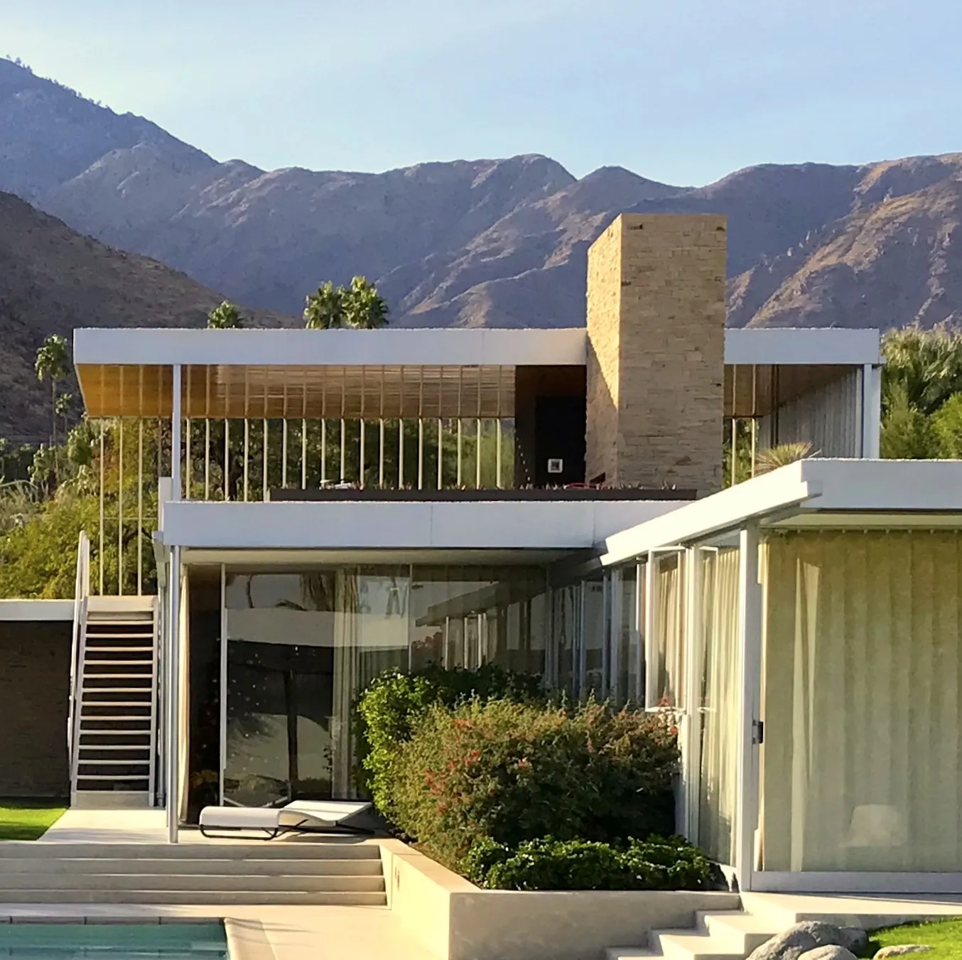 Close view of the Kaufmann Desert House showing the stone chimney, a white fascia above vertical slats, and a curtained glass wall with mountains behind.