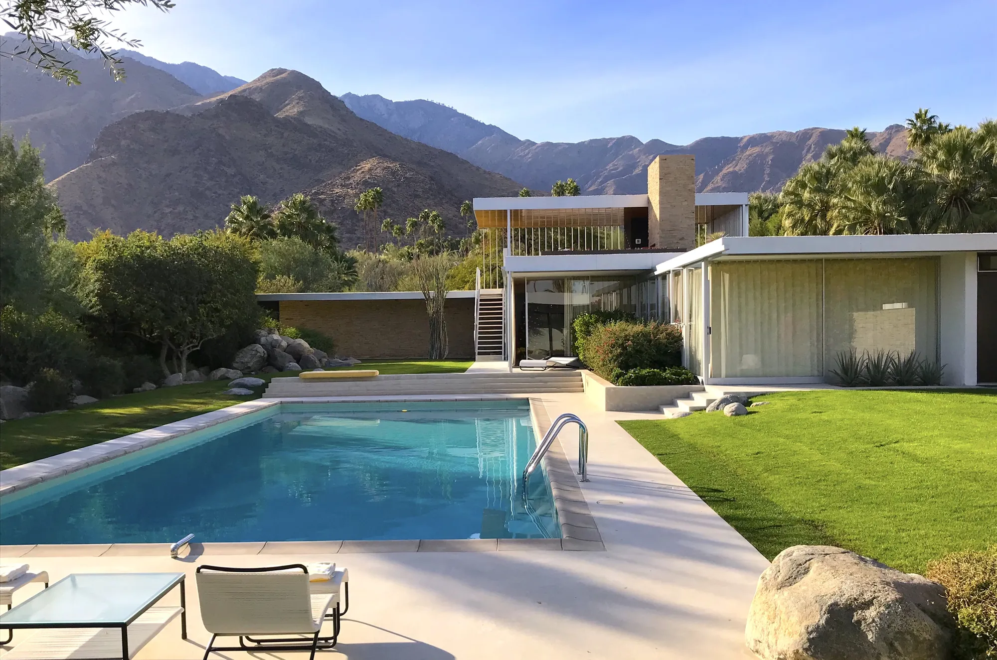 The Kaufmann Desert House seen across its swimming pool, with a stone chimney, a glass wall with curtains, a lawn and mountains behind.