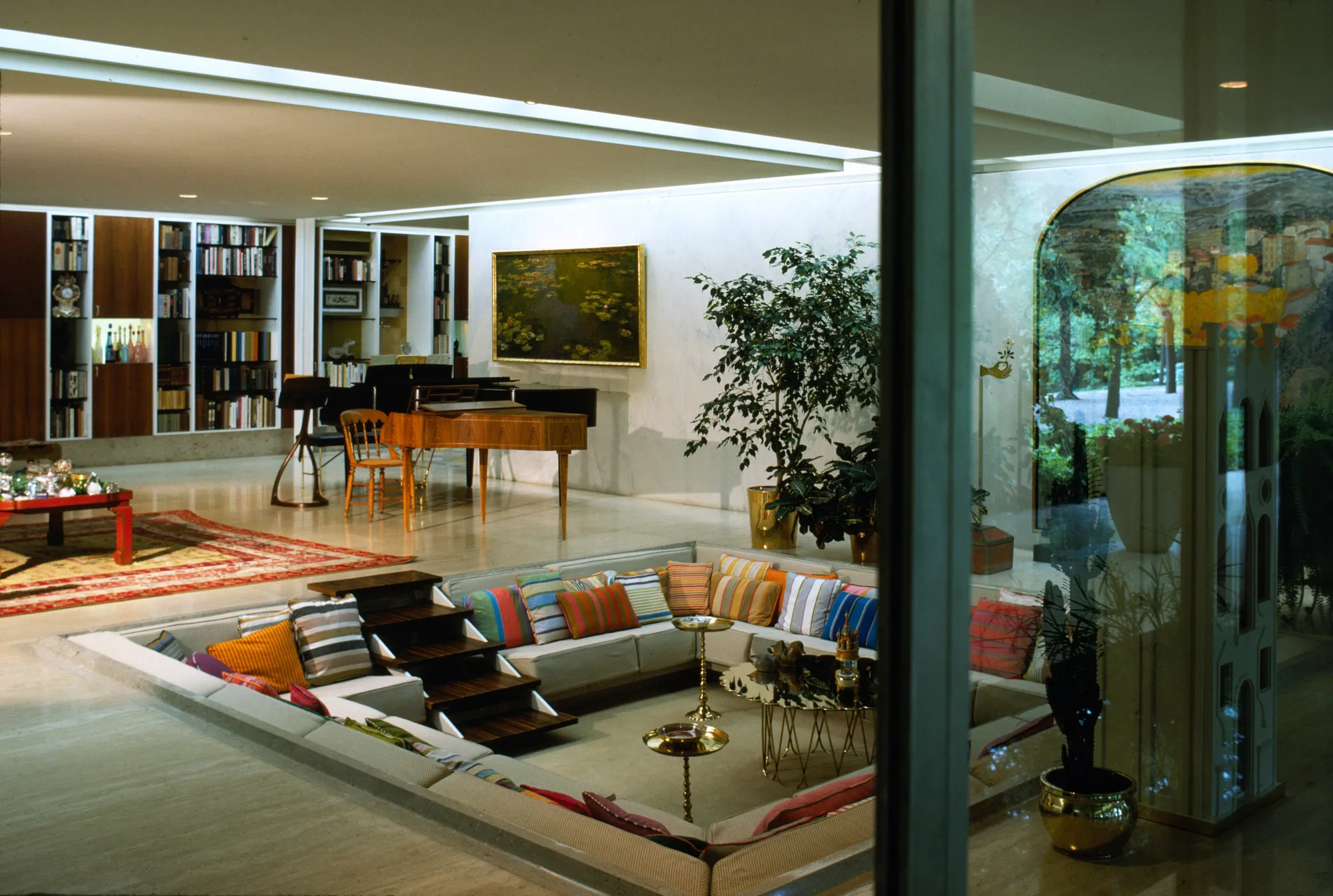 A sunken square seating area with striped cushions, a piano and bookshelves, seen through a glass wall.