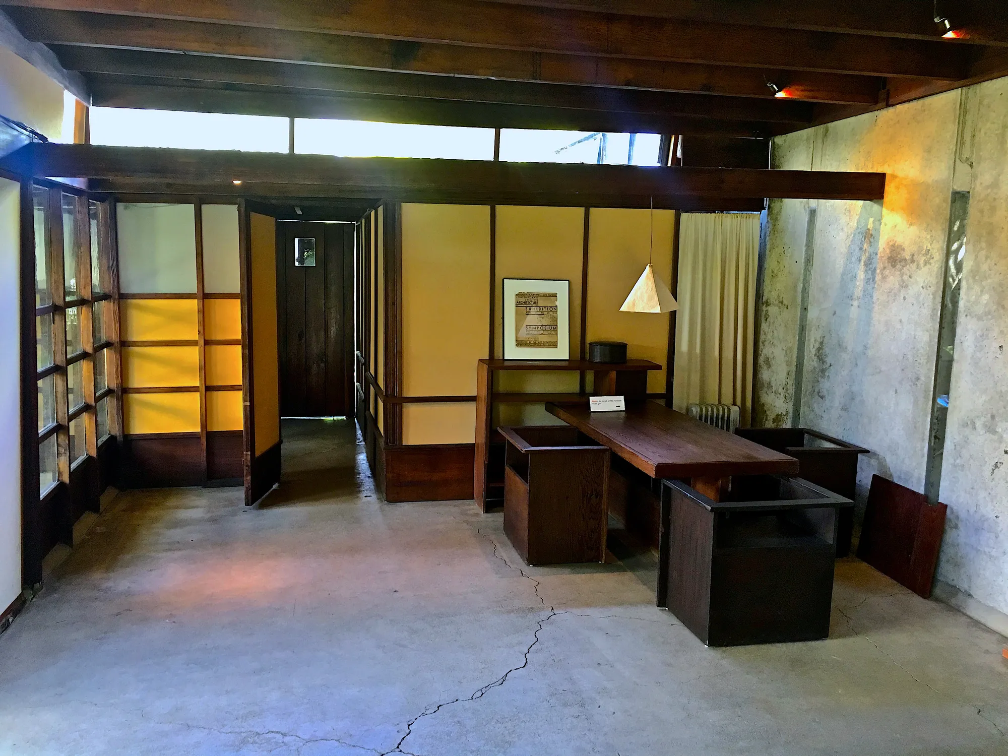 A room with a concrete floor, a rough concrete wall, wood-framed glass and panel screens, and dark wood furniture.